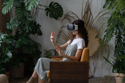 Young woman wearing vr helmet experiencing virtual reality while resting in greenhouse