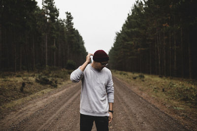 Young man touching knit hat while standing on dirt road amidst trees