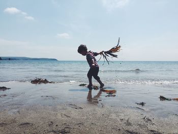 Boy with seaweed playing on shore at beach against sky