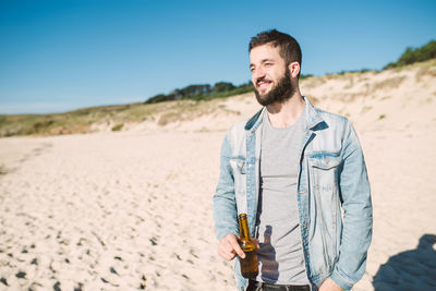 Young man holding alcoholic drink bottle while standing at beach during sunny day
