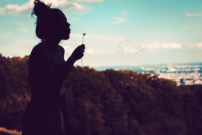 Side view of silhouette woman blowing dandelion against sky