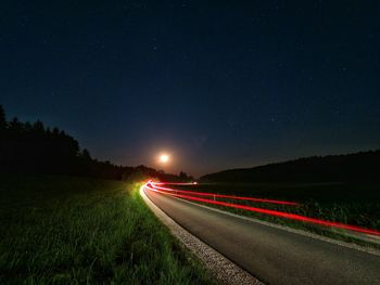 Light trails on road amidst field against sky at night