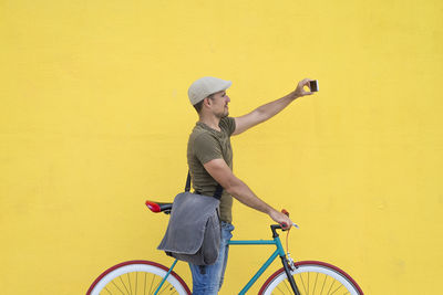 Man taking selfie while standing with bicycle against yellow wall