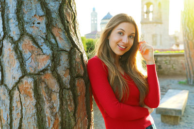 Portrait of smiling young woman standing by tree trunk