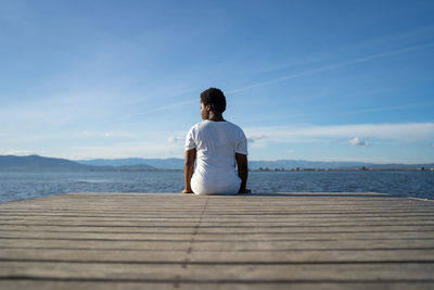 Rear view of man sitting on wood looking at sea against sky