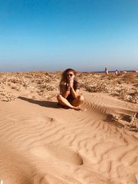 Full length of young woman sitting on sand at beach against clear sky