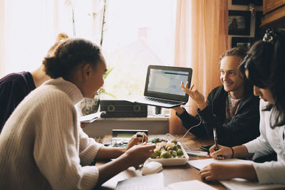 Smiling teenage boy showing laptop to friends while studying at table