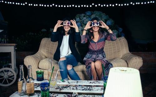 Portrait of young friends holding smart phones on eyes while on sofa during party