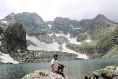 Man sitting on rock by lake against mountain