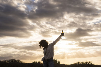 Woman holding bottle while standing against sky during sunset