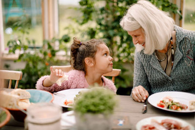 Smiling grandmother looking at granddaughter while sitting by dining table during lunch
