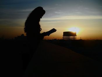 Side view of silhouette woman reading book against sky during sunset