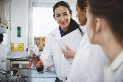 Confident mature teacher explaining to young female college students in chemistry laboratory