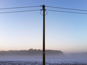 Low angle view of power line by snow covered field against sky