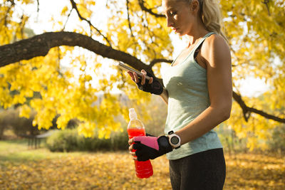 Side view of mid adult woman with bottle using smart phone in park during autumn