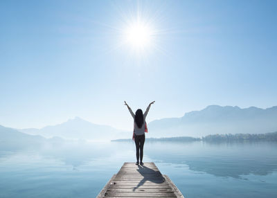 Full length rear view of woman with arms raised standing on pier by lake against sky during sunny day