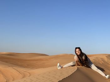 Portrait of woman sitting on sand dune in desert against clear blue sky