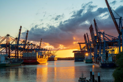Cranes at commercial dock against sky during sunset