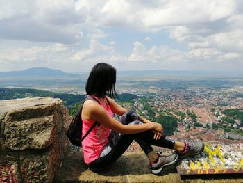 Womam sitting on a rock looking at cityscape against sky