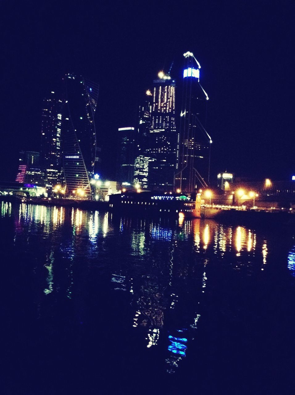 night, illuminated, city, water, building exterior, architecture, built structure, reflection, river, cityscape, skyscraper, clear sky, modern, waterfront, sky, city life, ferris wheel, urban skyline, dark, light - natural phenomenon