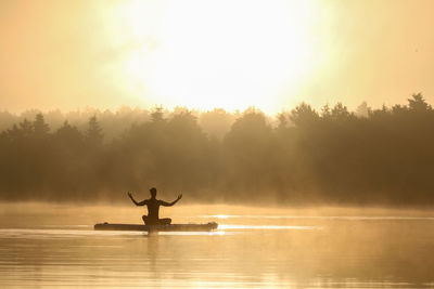 Silhouette woman meditating in lake against sky during sunrise