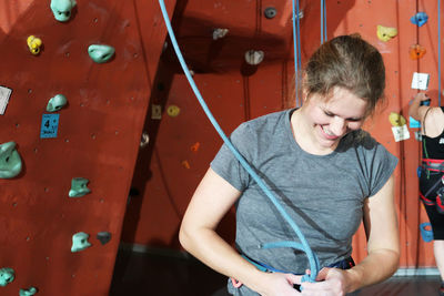 Smiling woman tying rope while standing against climbing wall