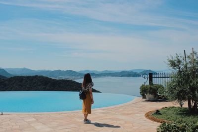 Rear view of woman standing by swimming pool against sky