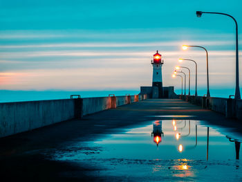 Illuminated street light by sea against sky during sunset