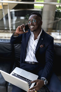 Smiling young male entrepreneur talking on smart phone with laptop in hotel lobby