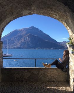 Woman sitting on shore by sea against mountains