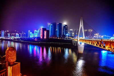 Illuminated bridge over river by buildings against sky at night
