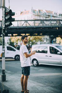 Side view of young man holding mobile phone while standing on sidewalk in city