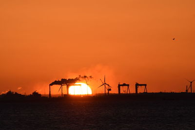 Silhouette of factory by sea against orange sky