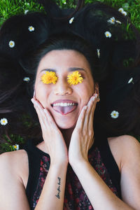 Top view of content female with tattoo and blooming flowers on eyes touching face while lying with tongue out on meadow