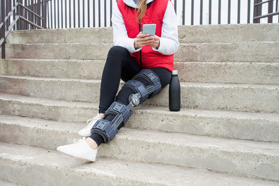 Woman wearing knee brace or orthosis after leg surgery, sitting on stairs ourdoors