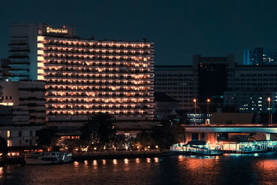 Illuminated buildings by river against sky at night