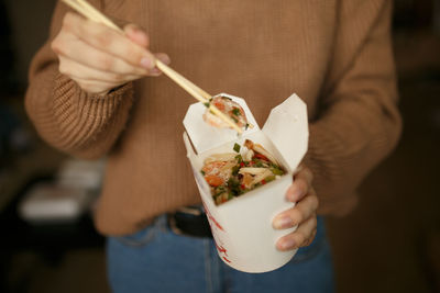 Girl in casual clothes eating chinese noodles from a box sitting on the floor of home, food delivery