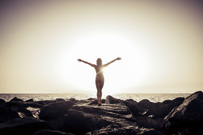 Rear view of woman with arms outstretched on rocky shore against clear sky