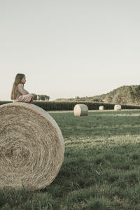 Child sitting on a hay bales on field against clear sky