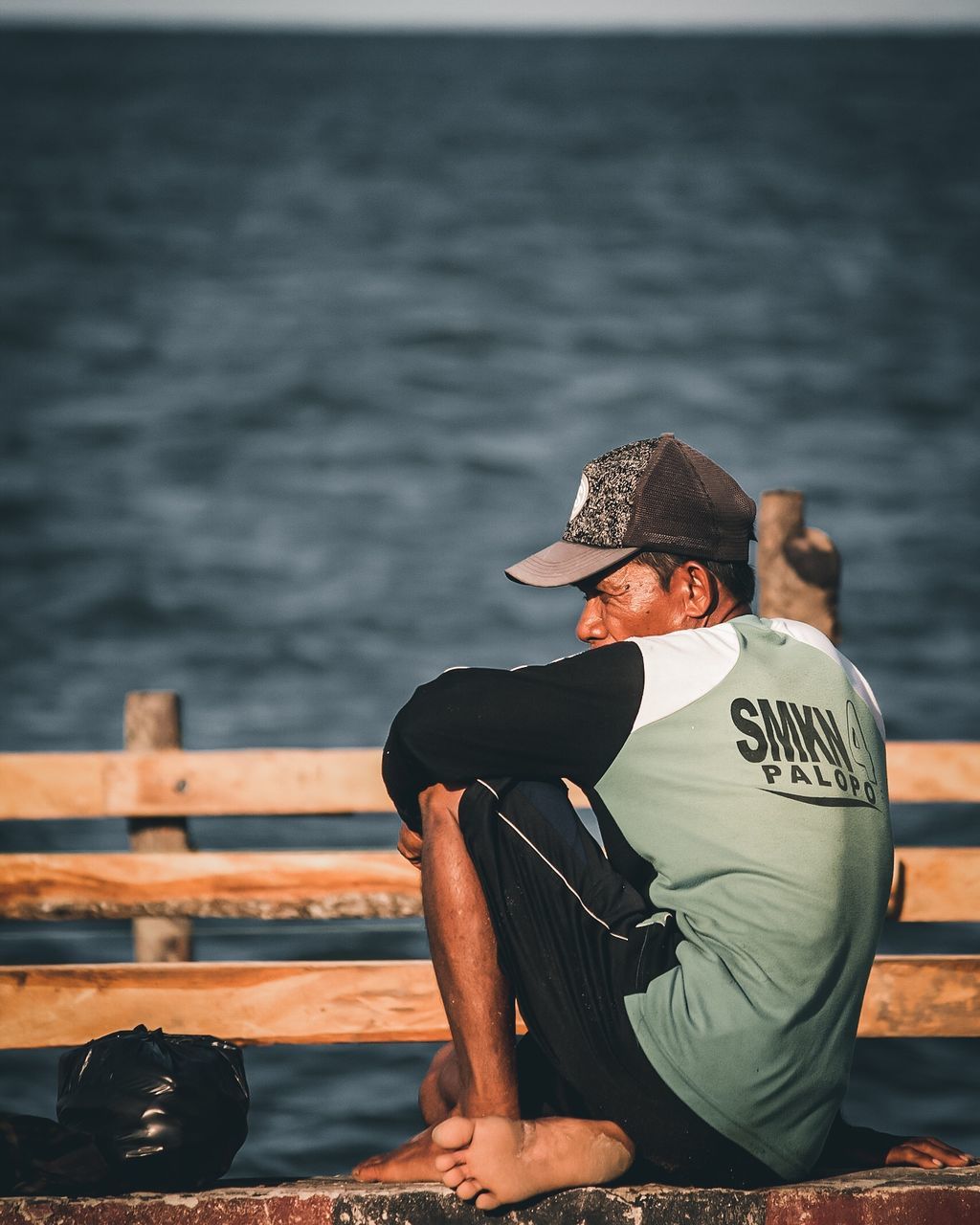 water, adult, sea, one person, nature, clothing, sitting, men, relaxation, lifestyles, hat, full length, leisure activity, outdoors, day, young adult, land, beach, sky, casual clothing, nautical vessel, black, pier, person, copy space, tranquility, cap