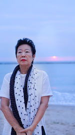 Thoughtful senior woman standing at beach against sky during sunset