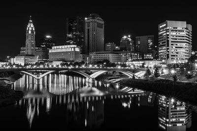 Illuminated modern buildings by river against sky at night