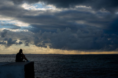 Man sitting by sea against sky during sunset