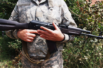 Midsection of military man holding rifle against plants