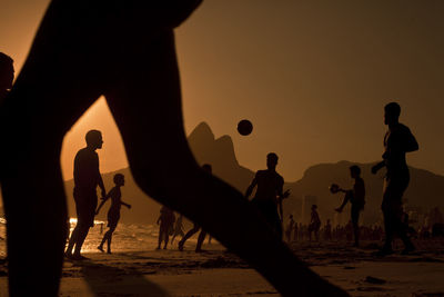 Silhouette friends playing soccer at beach against orange sky during sunset