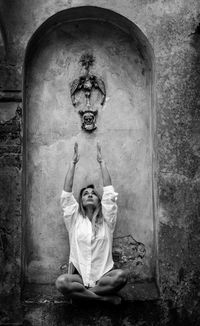 Full length of woman with arms raised sitting by wall