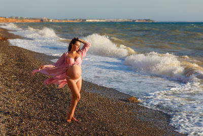 Beautiful pregnant woman standing on beach in light dress in the wind. pleasure