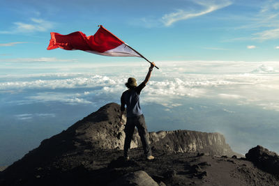 Rear view of man standing on rock with indonesian flag against sky