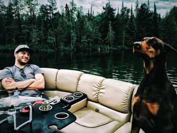 Happy man on sofa looking at doberman pinscher by lake in forest