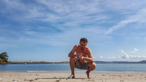 Full length of shirtless man crouching at beach against sky